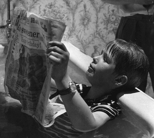 Black and white photo of a young boy reading a newspaper in a bathtub, fully clothed