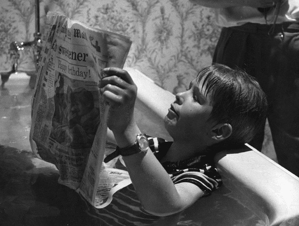 Black and white photo of a young boy reading a newspaper in a bathtub, fully clothed