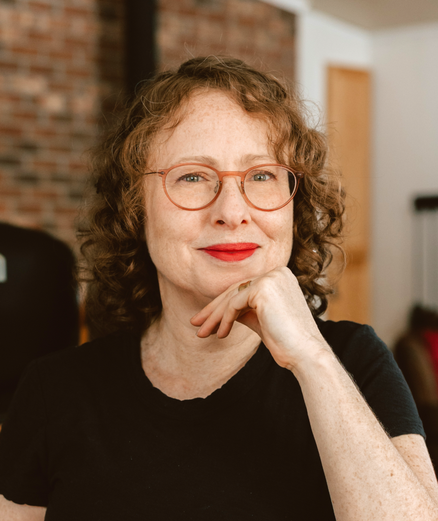 Janice, age 56, wearing glasses, a black t-shirt, and pink lipstick, smiling