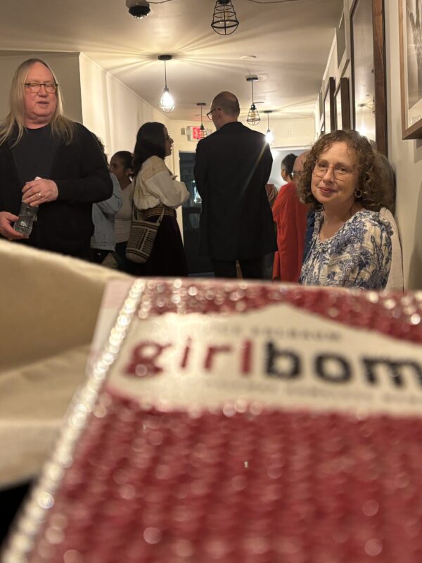 A copy of Girlbomb with a bedazzled cover in the foreground, Janice and party guests in the background